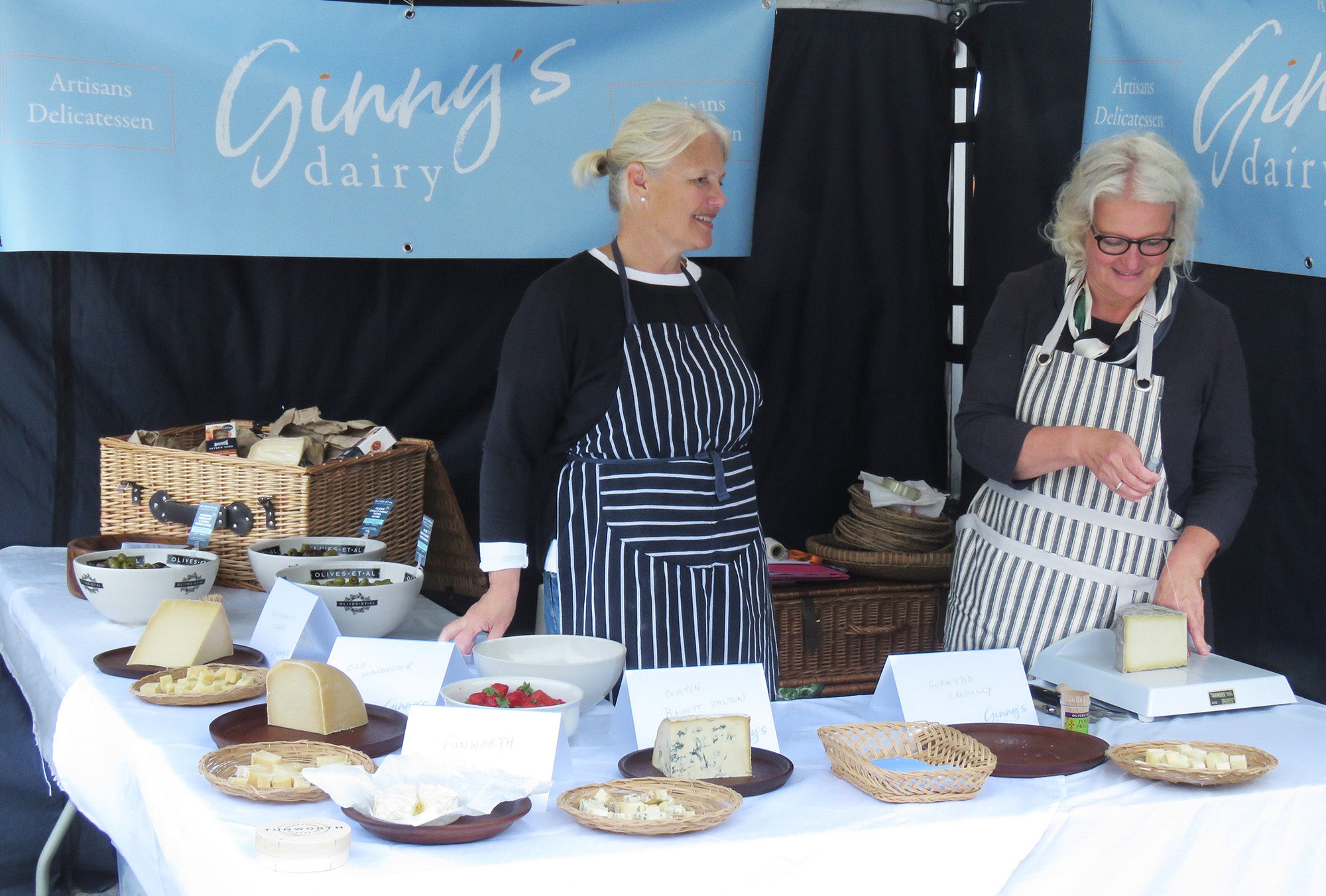 Jane and Sarah who run Ginny's Dairy cutting cheese at a market stall covered in plates of cheese