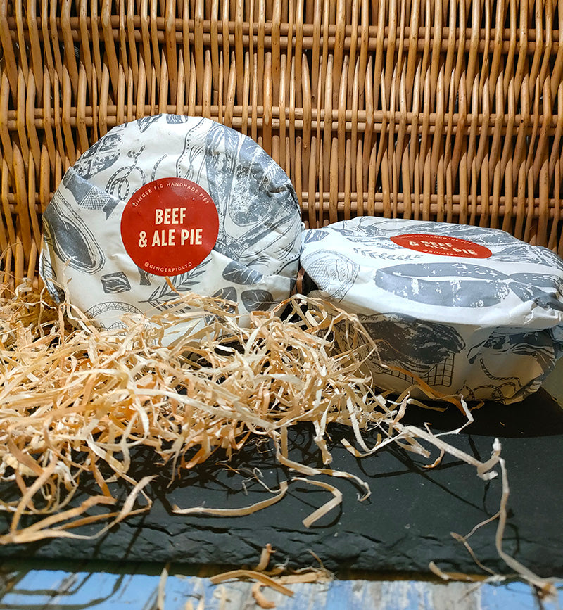 Two beef and ale pies lie on a slate before a wicker backdrop with straw in the foreground