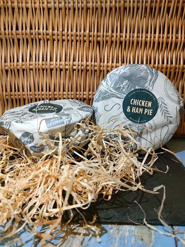 Two chicken and ham pies sit on slate before a wicker backdrop with straw in the foreground