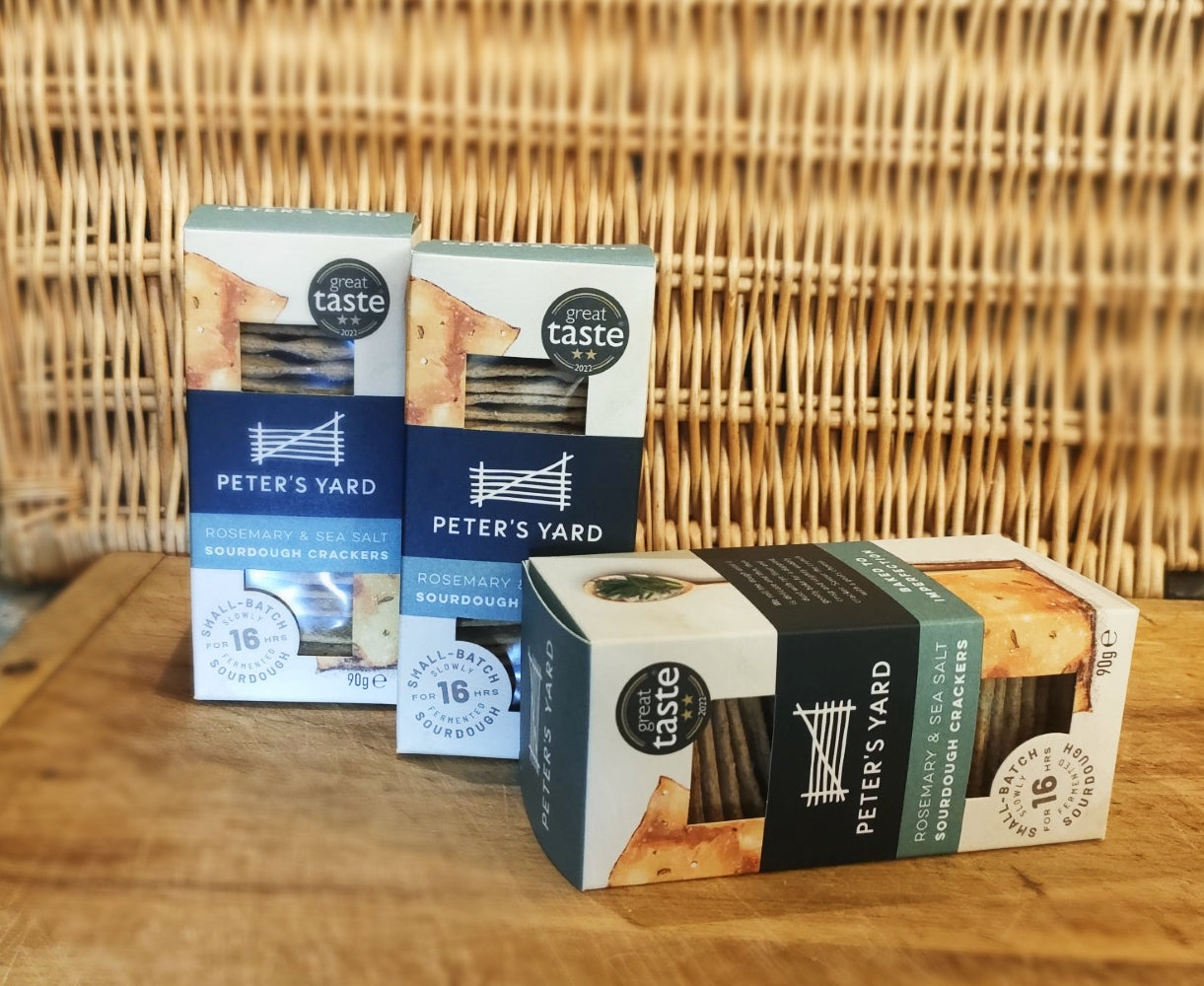 Rosemary and sea salt crackers in three boxes, on a wooden cheeseboard. Sat in front of a picnic hamper