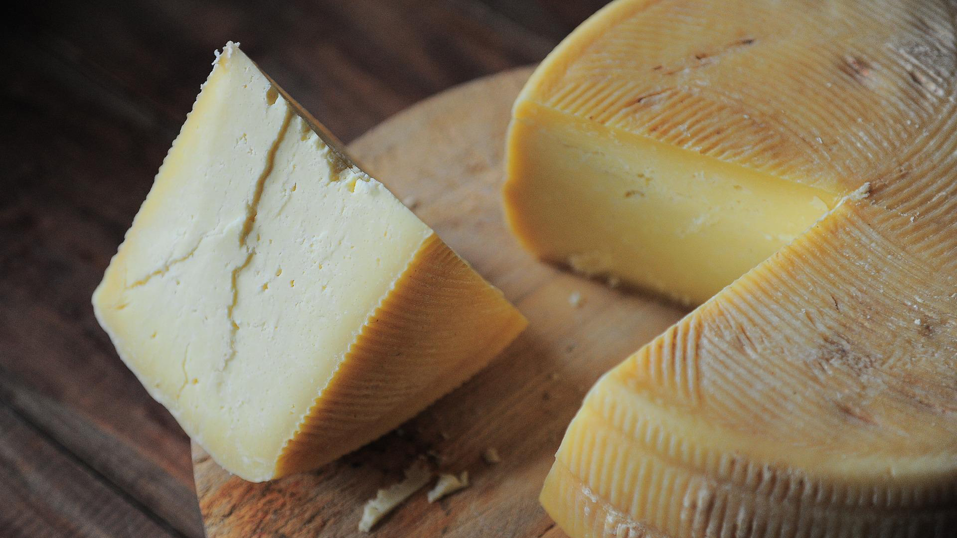 A chunk of cheese cut from a round slab of cheese on a wooden chopping board