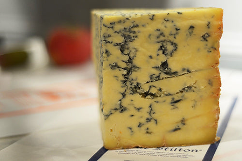 A large piece of Colston Bassett Stilton sits on a piece of paper with the name printed on it. It is yellow tinged with dark mould ripples throughout it.