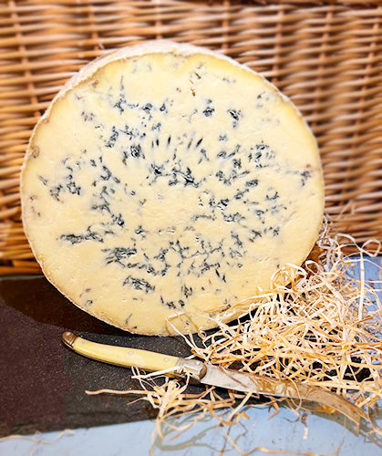 A wheel of Colston Bassett Stilton cheese stands on its side on a slate with a wicker background, with straw and a knife before it