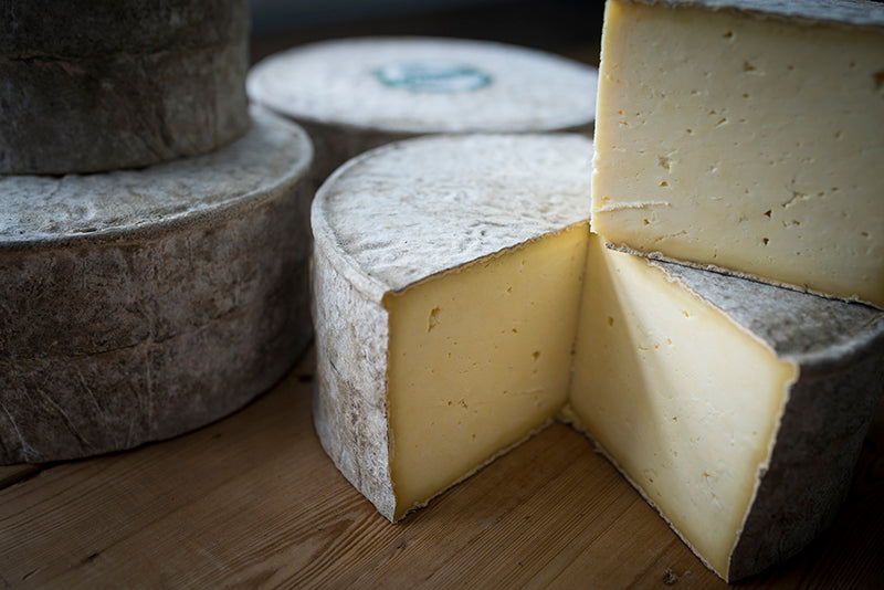 Large wheels of Gorwydd Caerphilly cheese sit on top of each other on a wooden table - one has a quarter of cheese cut out of it.