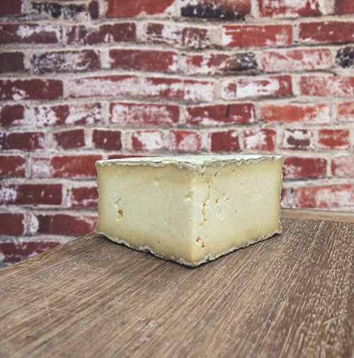 A large section of Gorwydd Caerphilly cheese sits on the corner of a wooden table with a red brick wall in the background. The cheese is pale yellow and dapples with some holes.