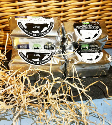 Two stacks of butter wrapped in silver packaging - Meggy Moo's Sea Salted butter and Meggy Moo's Peppered Butter - on a slate with a wicker background, surrounded by straw