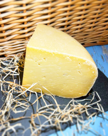 A quarter of a wheel of Old Winchester cheese on a piece of slate with a wicker background, surrounded by straw